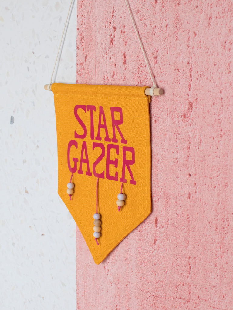 A yellow canvas banner that reads Star Gazer and is decorated with hanging beads is hung on a white and pink textured wall.