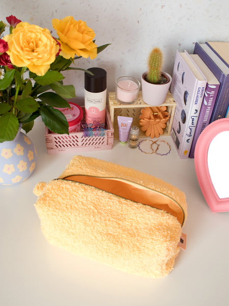 A yellow towelling zip pouch opened on a busy dressing table filled with beauty products, books and a vase of flowers.