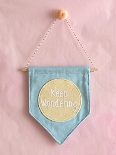 A blue canvas banner that reads 'Keep Wondering' in a yellow circle is hung on a pink wall.