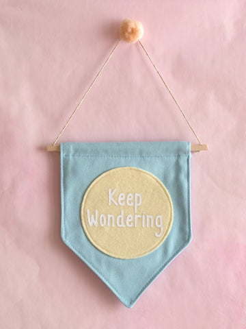 A blue canvas banner that reads 'Keep Wondering' in a yellow circle is hung on a pink wall.