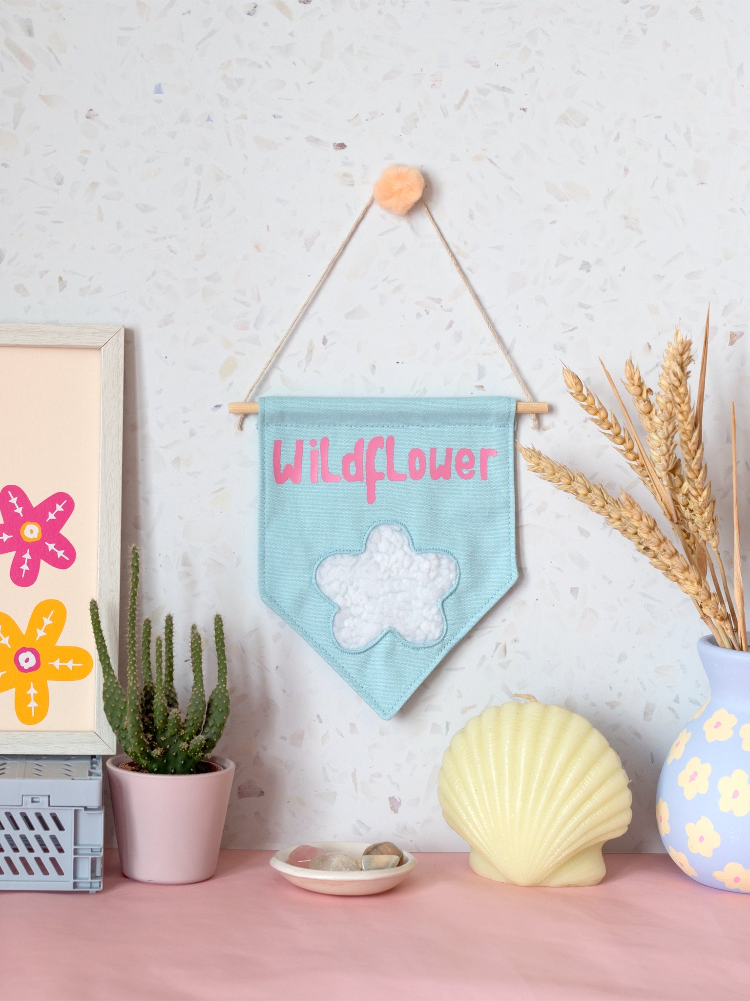 A blue canvas banner that reads Wildflower with a fluffy white flower at the bottom is hung on a white terrazzo wall surrounded by framed art, a cactus,  flowers and a shell candle.