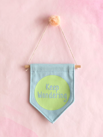 A blue canvas banner that reads 'Keep Wondering' in a green circle is hung on a pink wall.
