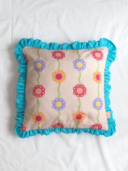 A pink floral printed cushion with blue ruffles in the centre of a white bedsheet.