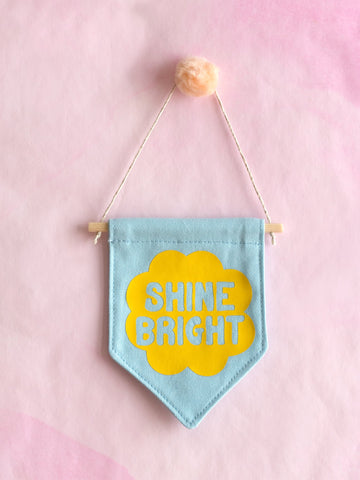 A blue canvas banner that reads Shine Bright in a yellow flower is hung on a pink wall.