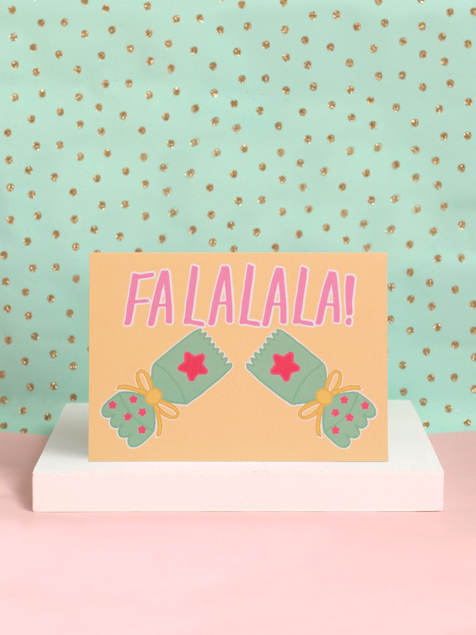 A landscape Christmas card with Falalala and a popped cracker on the front sat on a white block.