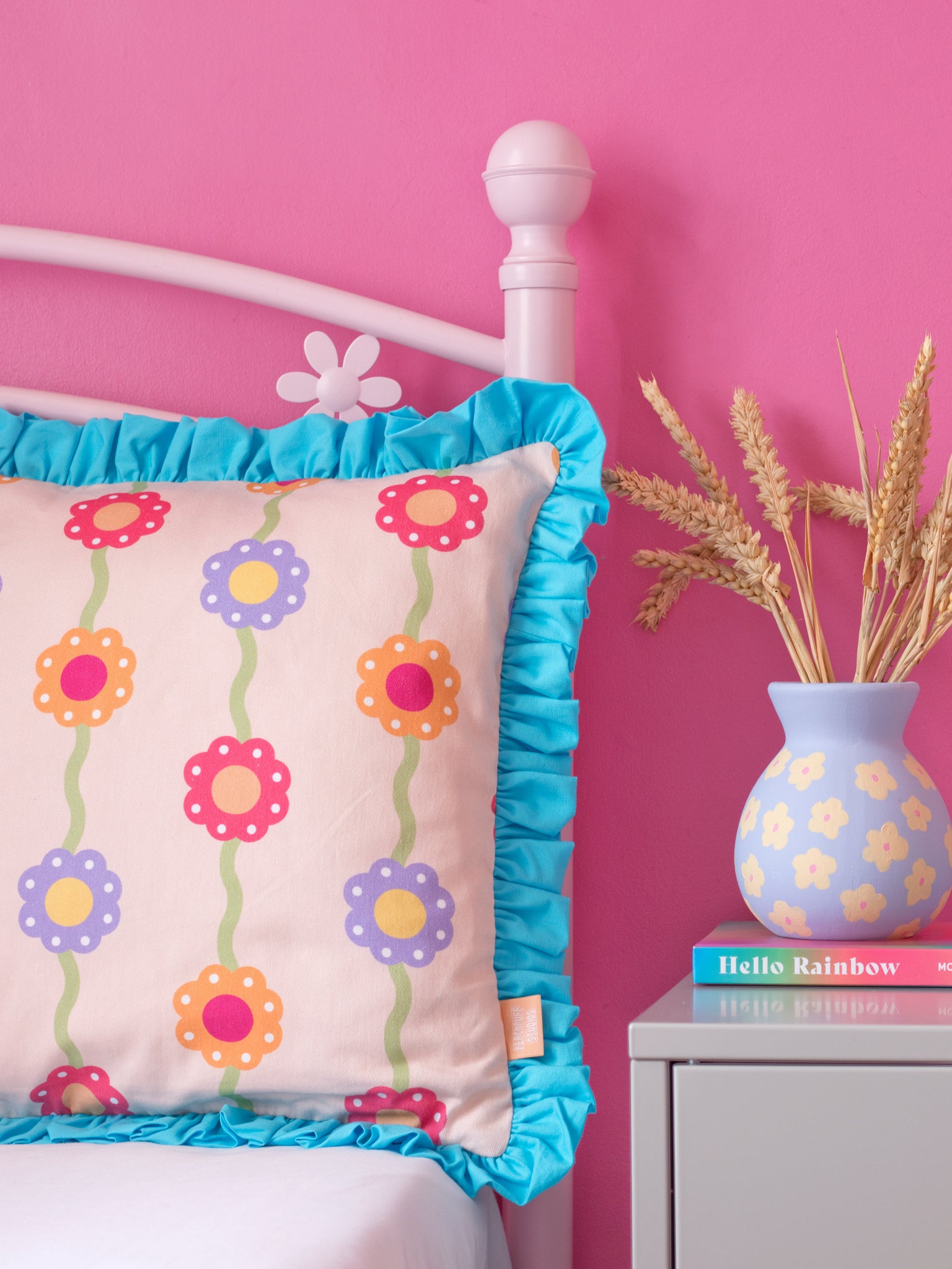 A pink floral printed cushion with blue ruffles on a pink flower bed next to a dressing table styled with dried flowers and a book.