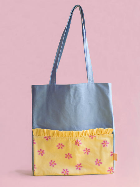 A blue tote bag with a yellow front pocket stands against a pink foreground.