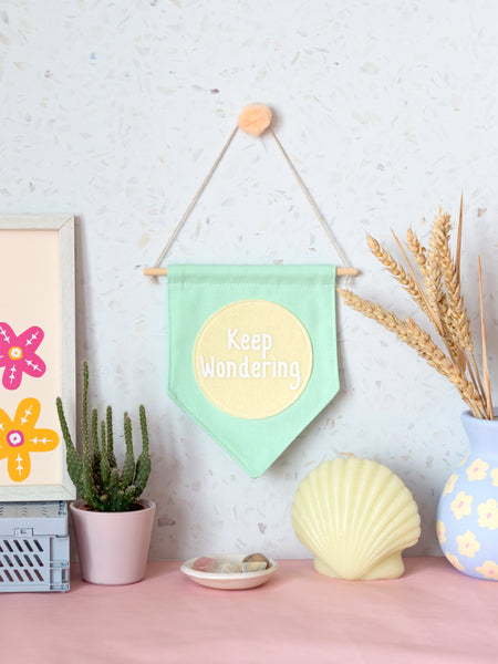 A green canvas banner that reads 'Keep Wondering' in a yellow circle is hung on a white terrazzo wall surrounded by framed art, a cactus,  flowers and a shell candle.