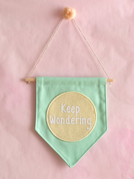 A green canvas banner that reads 'Keep Wondering' in a yellow circle is hung on a pink wall.