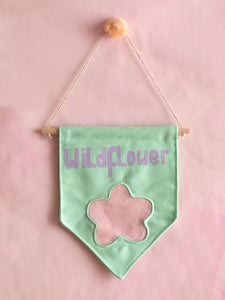 A green canvas banner that reads Wildflower with a fluffy pink flower at the bottom is hung on a pink wall.