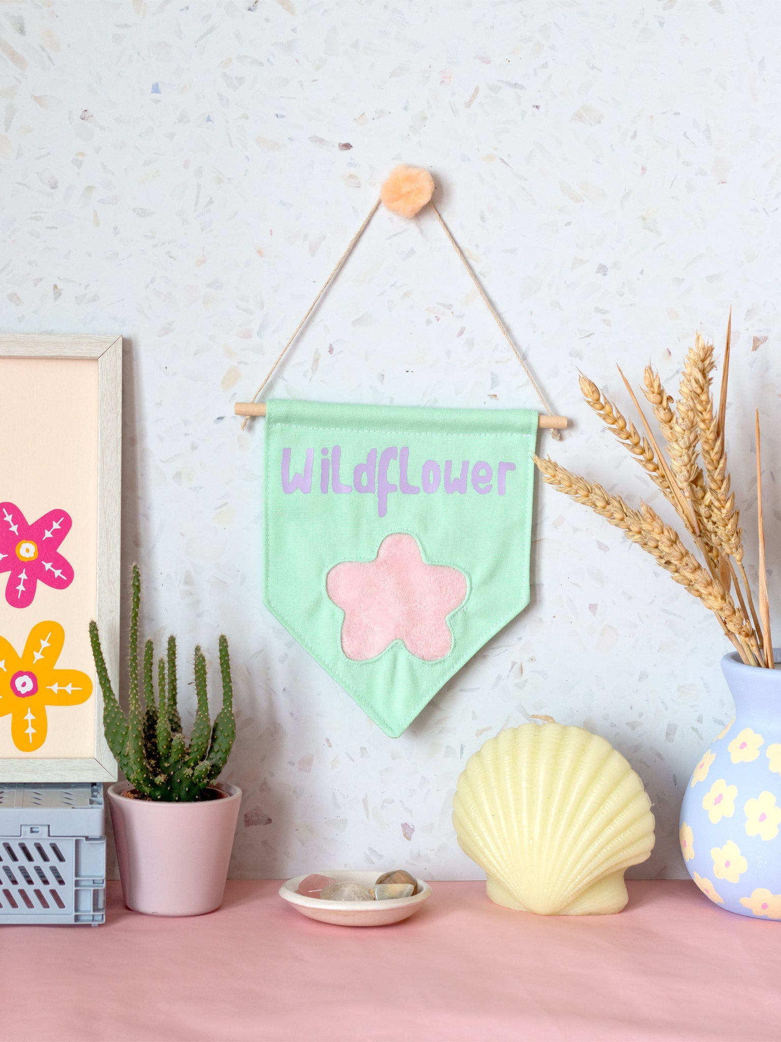 A green canvas banner that reads Wildflower with a fluffy pink flower at the bottom is hung on a white terrazzo wall surrounded by framed art, a cactus,  flowers and a shell candle.