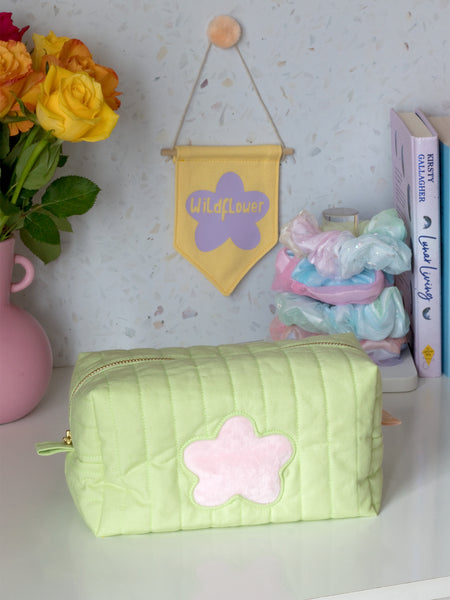A green quilted zip pouch with a fluffy pink flower in the centre on a dressing table filled with a yellow wall hanging, scrunchies, books, and a vase of flowers.