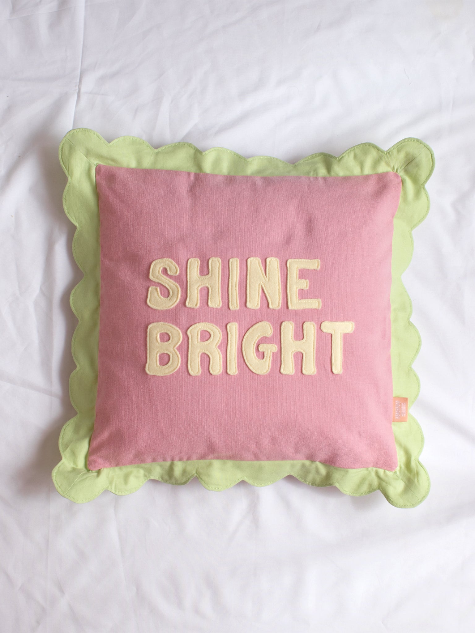 A pink cushion with green scalloped trim and 'Shine Bright' lettering on the front in the centre of a white bedsheet.