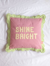 A pink cushion with green scalloped trim and 'Shine Bright' lettering on the front in the centre of a white bedsheet.