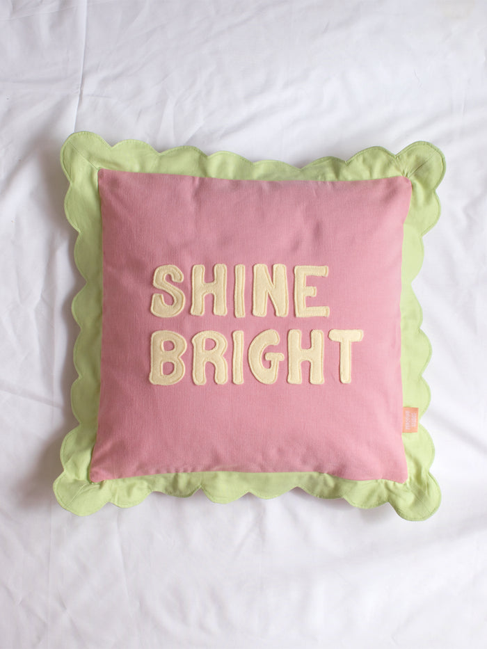 A pink cushion with green scalloped trim and 'Shine Bright' lettering on the front in the centre of a white bedsheet.
