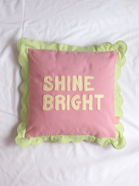 A pink cushion with green scalloped trim and 'Shine Bright' lettering on the front in the centre of a white bedsheet.