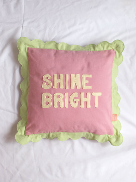 A pink cushion with green scalloped trim and 'Shine Bright' lettering on the front in the centre of a white bedsheet.