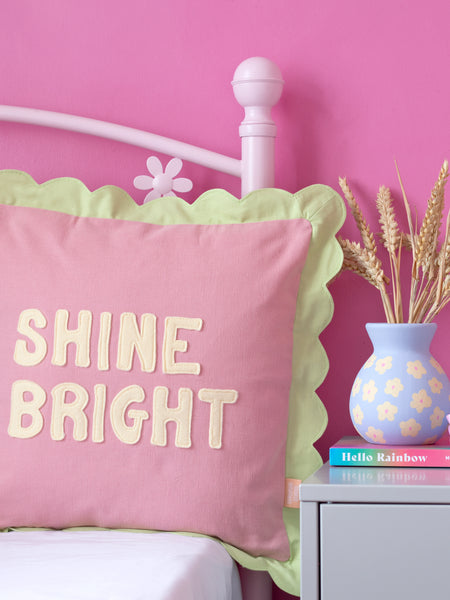 A pink cushion with green scalloped trim and 'Shine Bright' lettering on the front on a pink flower bed next to a dressing table styled with dried flowers and a book.