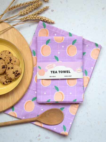 A kitchen style flat lay with a lilac tea towel with peaches printed on, wrapped in a creative belly band