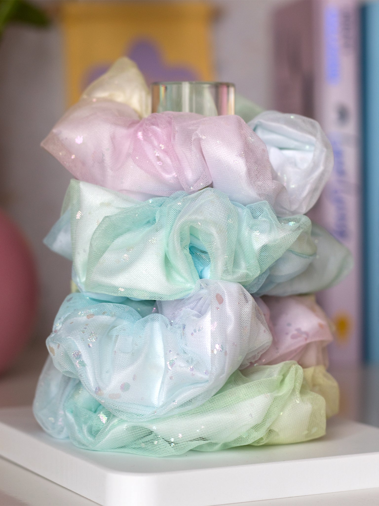 Four rainbow-coloured scrunchies are placed on top of each other on a white and clear holder.