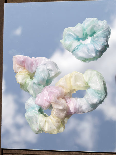 Four rainbow-coloured scrunchies are placed on a mirror where you can see the blue and cloudy reflection from the sky.