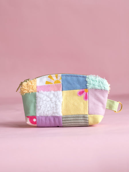 A multi-coloured patchwork coin purse with a metal zip and ring hook on a pink foreground.