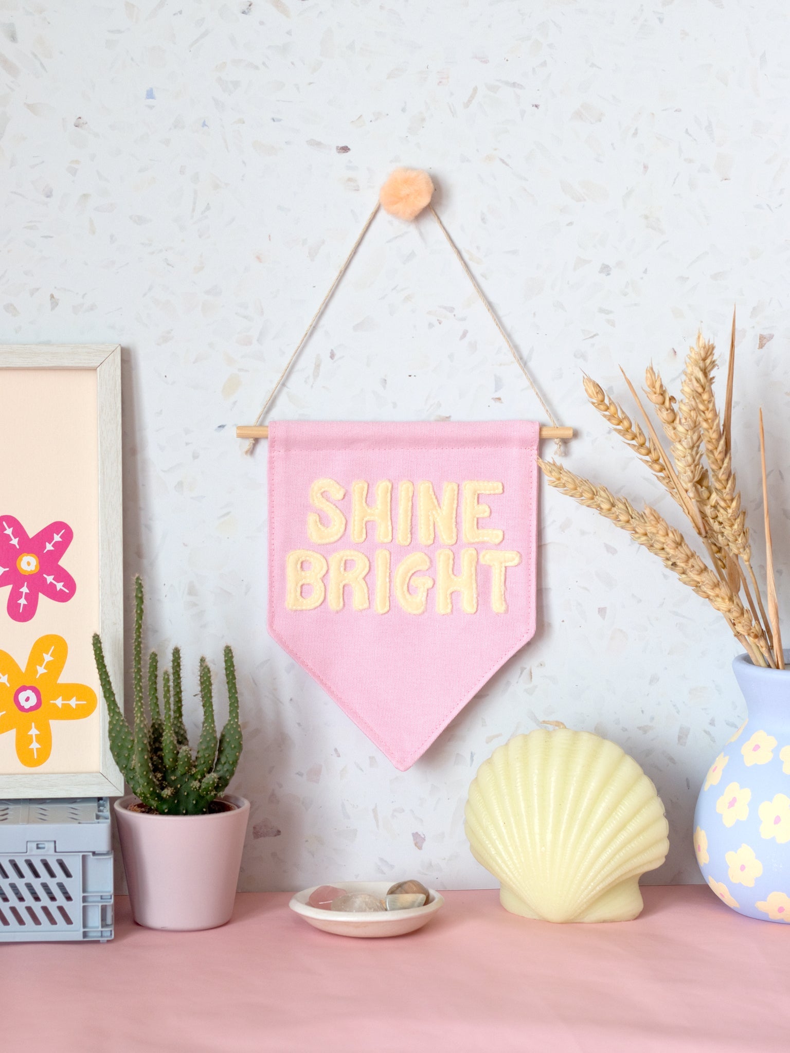 A pink canvas banner that reads Shine Bright in yellow fleece lettering is hung on a white terrazzo wall surrounded by framed art, a cactus,  flowers and a shell candle.