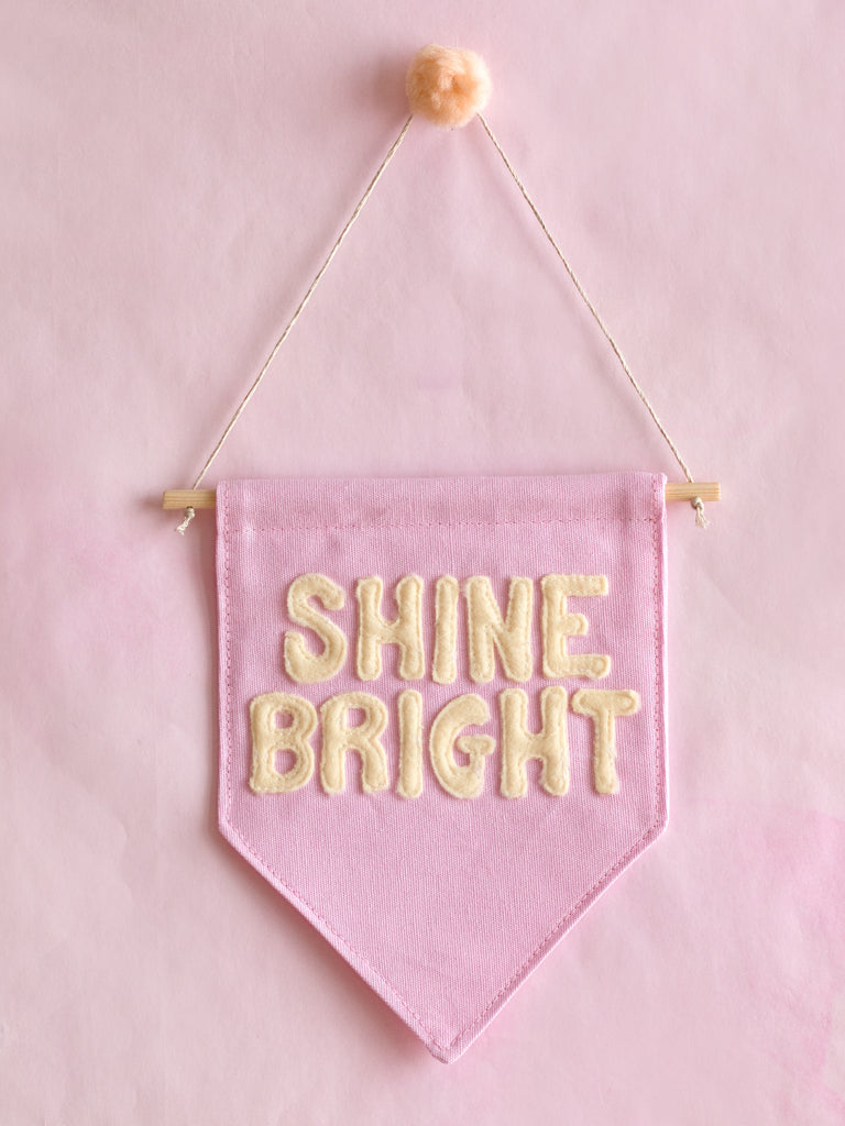 A pink canvas banner that reads Shine Bright in yellow fleece lettering is hung on a pink wall.
