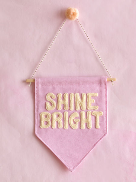 A pink canvas banner that reads Shine Bright in yellow fleece lettering is hung on a pink wall.