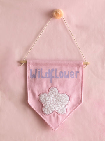 A pink canvas banner that reads Wildflower with a fluffy white flower at the bottom is hung on a pink wall.
