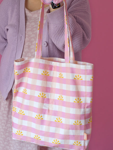 A pink gingham tote bag is held on the arm of a female wearing a purple patterned dress and cardigan.