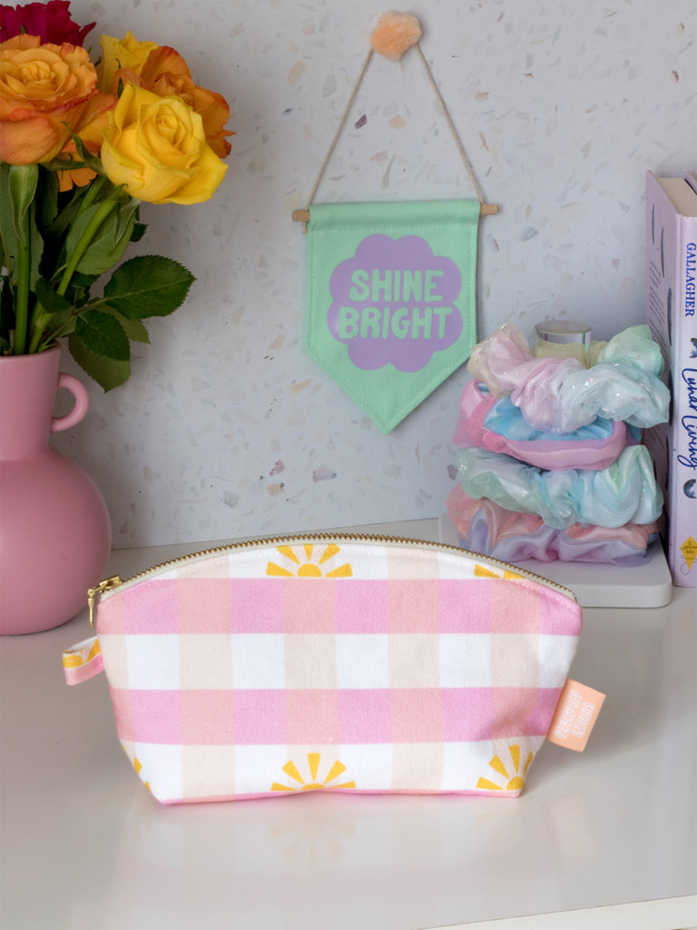 A pink gingham printed zip pouch in the centre on a dressing table filled with a green wall hanging, scrunchies, books, and a vase of flowers.
