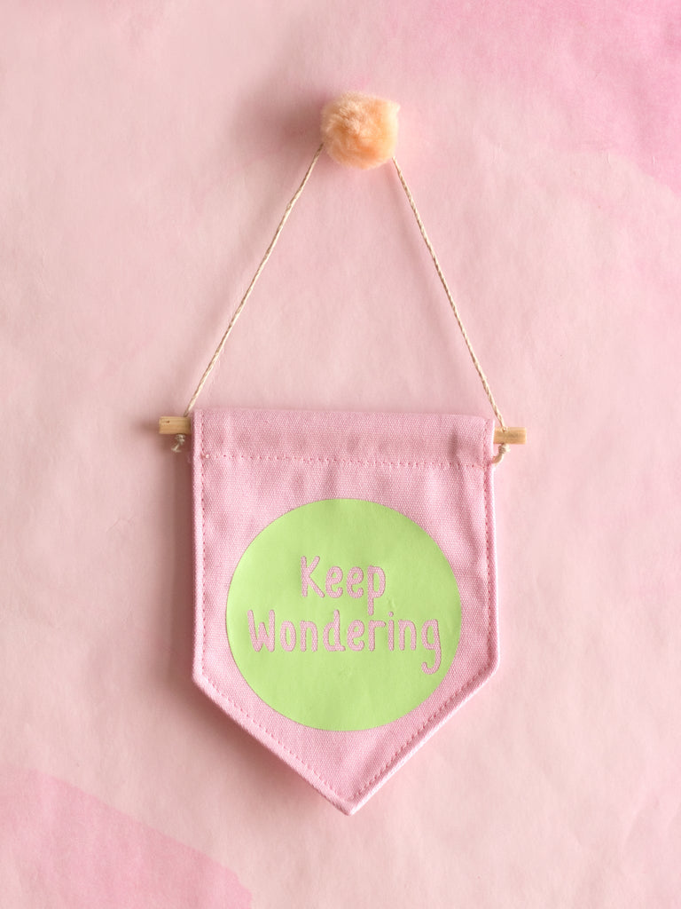 A pink canvas banner that reads 'Keep Wondering' in a green circle is hung on a pink wall.