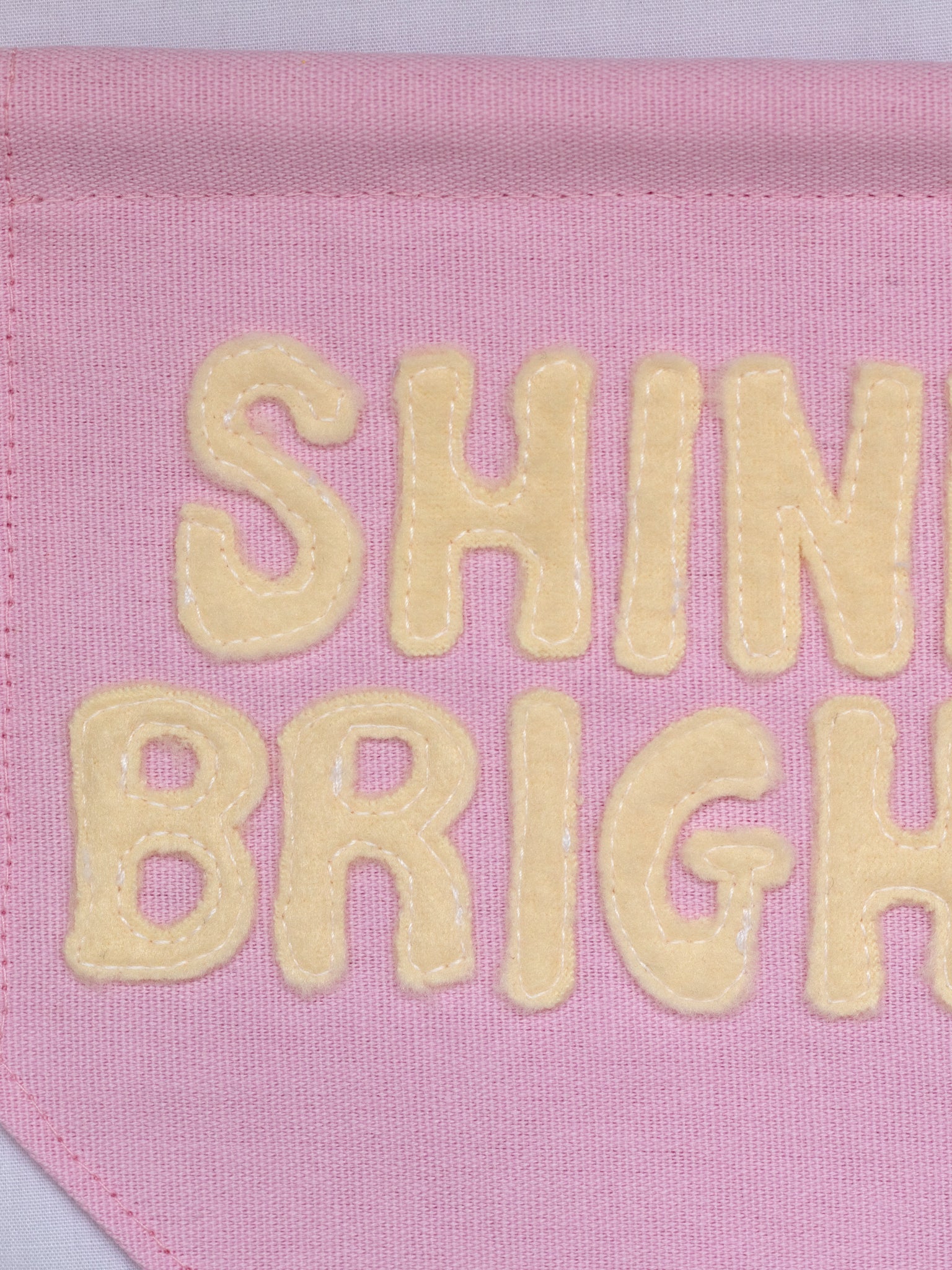 A close-up of a pink canvas banner that reads Shine Bright in yellow fleece lettering.