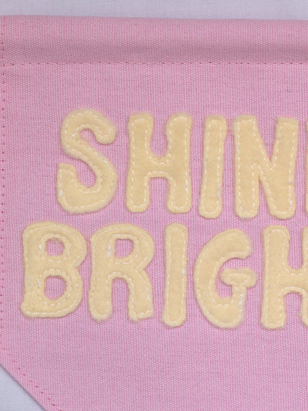 A close-up of a pink canvas banner that reads Shine Bright in yellow fleece lettering.