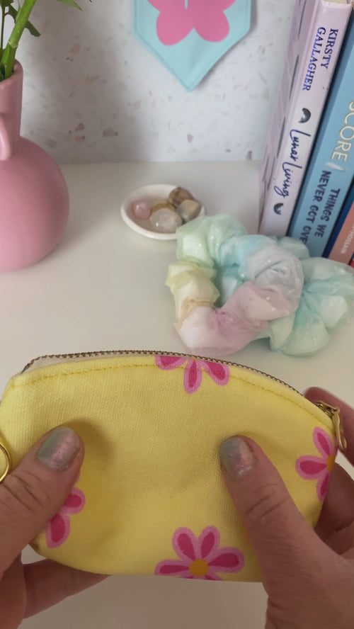 A pink gingham and yellow sunshine printed with a metal zip and ring hook is opened with the content placed inside, at the dressing table with books, scrunchies and a vase of flowers.