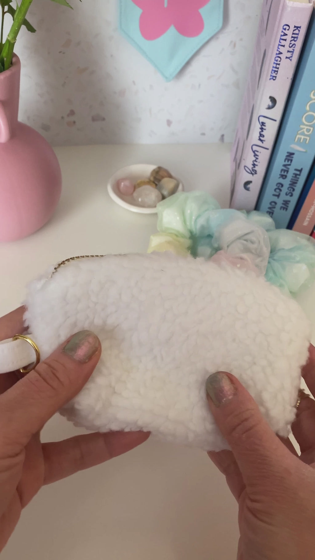 A white fluffy coin purse with a metal zip and ring hook is opened and shown at a dressing table with books, scrunchies and a vase of flowers.