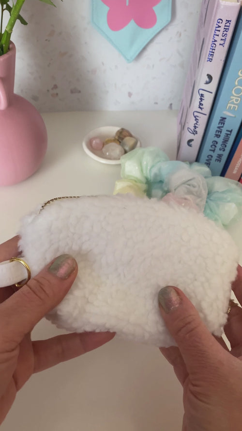 A white fluffy coin purse with a metal zip and ring hook is opened and shown at a dressing table with books, scrunchies and a vase of flowers.
