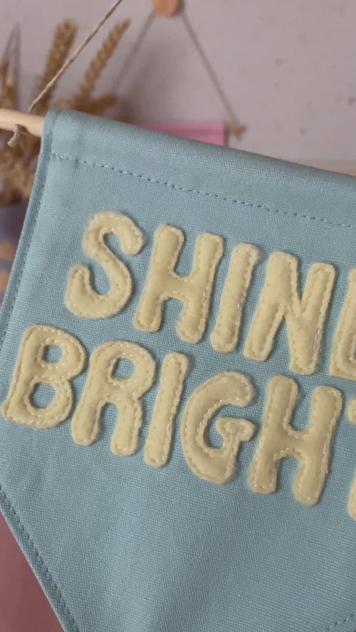A blue canvas banner that reads Shine Bright in yellow fleece lettering is shown to a camera.