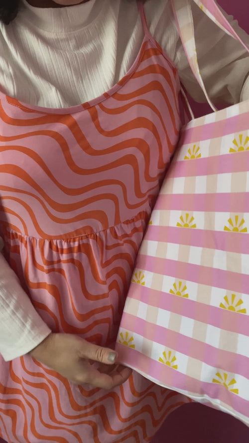 A pink gingham tote bag is worn on a shoulder of a female who opens the bag to reveal an inside zipped pocket.