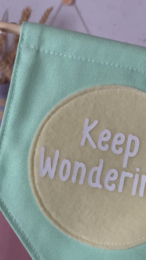 A green canvas banner that reads 'Keep Wondering' in a yellow circle is shown to a camera.