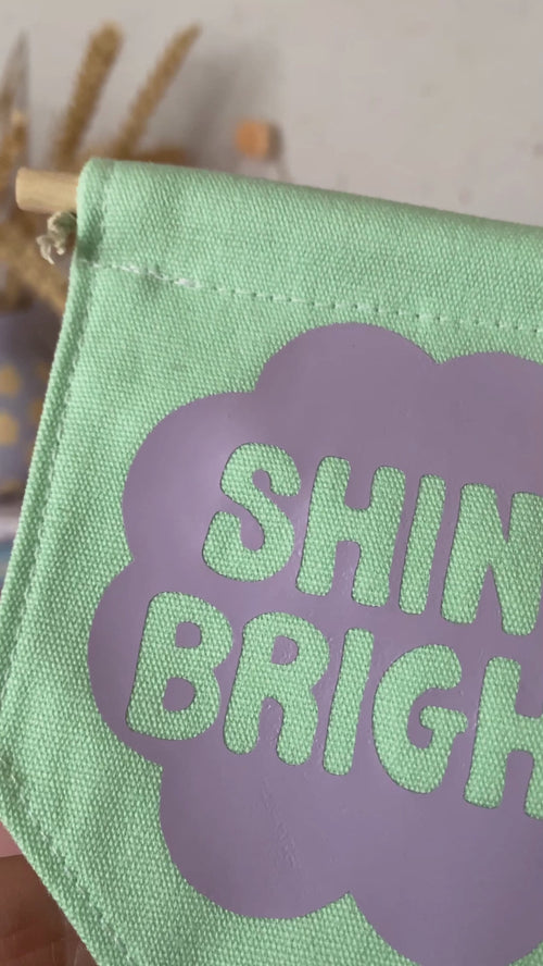 A green canvas banner that reads Shine Bright in a purple flower is shown to a camera.