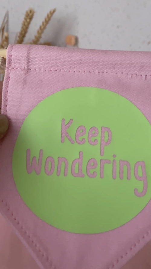A pink canvas banner that reads 'Keep Wondering' in a green circle is shown to a camera.