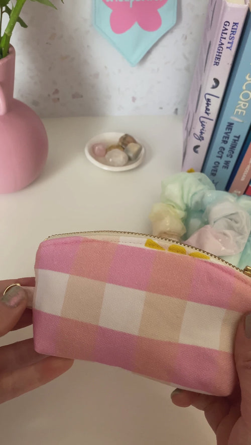 A pink gingham and yellow sunshine printed with a metal zip and ring hook is opened and shown the content at the dressing table with books, scrunchies and a vase of flowers.