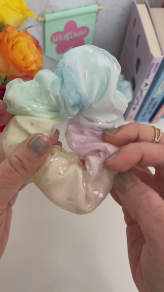 A rainbow scrunchie is shown to the camera while it's stretched open and closed. A female is wearing two scrunchies on her wrist, and she spins them back and forth.