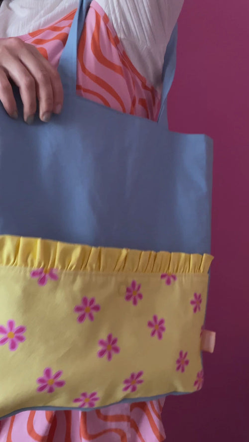 A blue tote bag with a yellow pocket is worn on a shoulder of a female who opens the bag to reveal an inside zipped pocket.