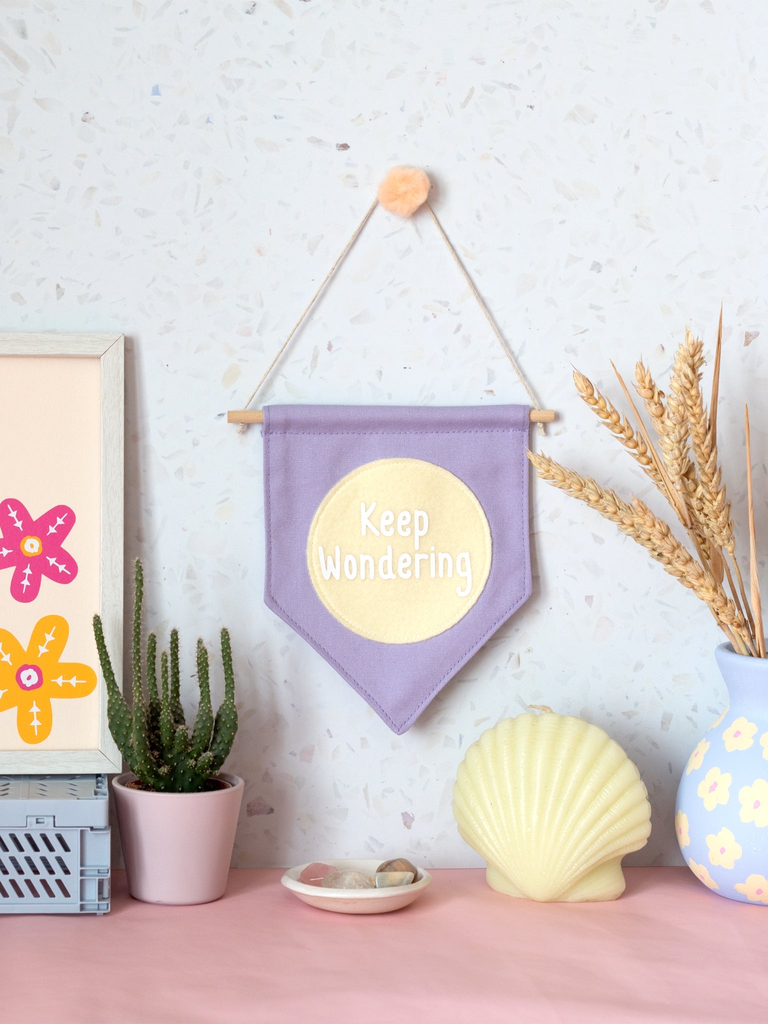 A purple canvas banner that reads 'Keep Wondering' in a yellow circle is hung on a white terrazzo wall surrounded by framed art, a cactus,  flowers and a shell candle.