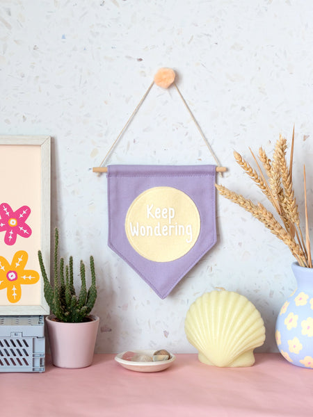 A purple canvas banner that reads 'Keep Wondering' in a yellow circle is hung on a white terrazzo wall surrounded by framed art, a cactus,  flowers and a shell candle.