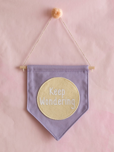 A purple canvas banner that reads 'Keep Wondering' in a yellow circle is hung on a pink wall.