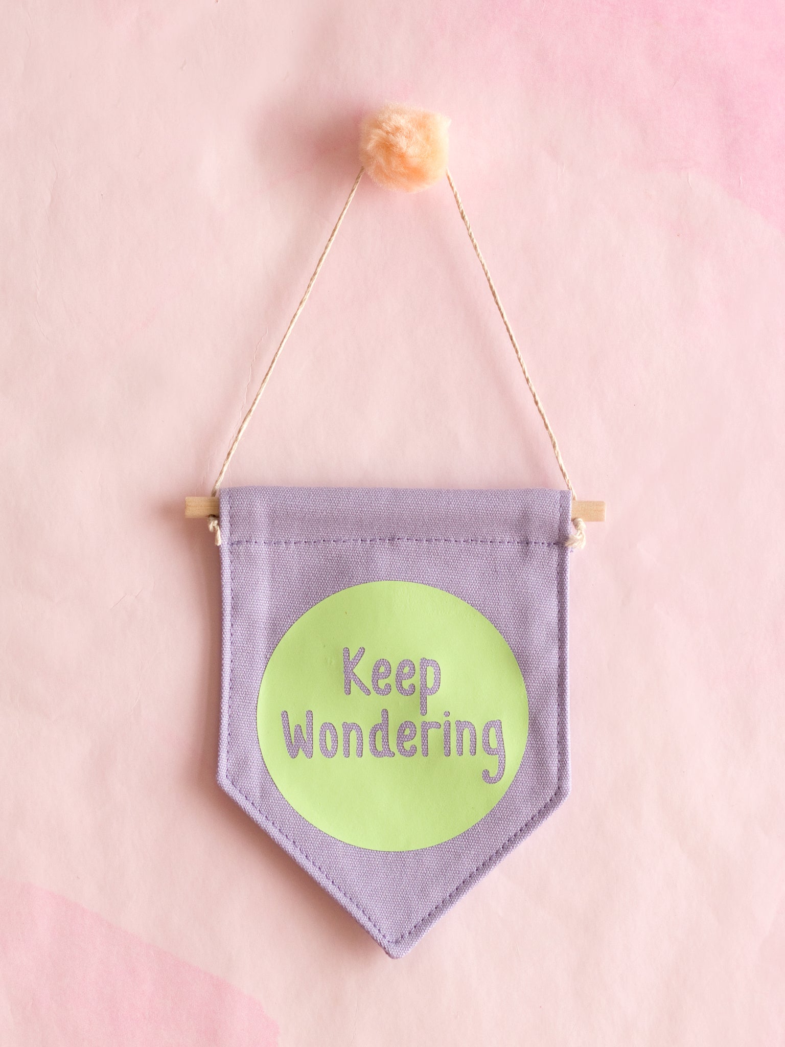 A purple canvas banner that reads 'Keep Wondering' in a green circle is hung on a pink wall.
